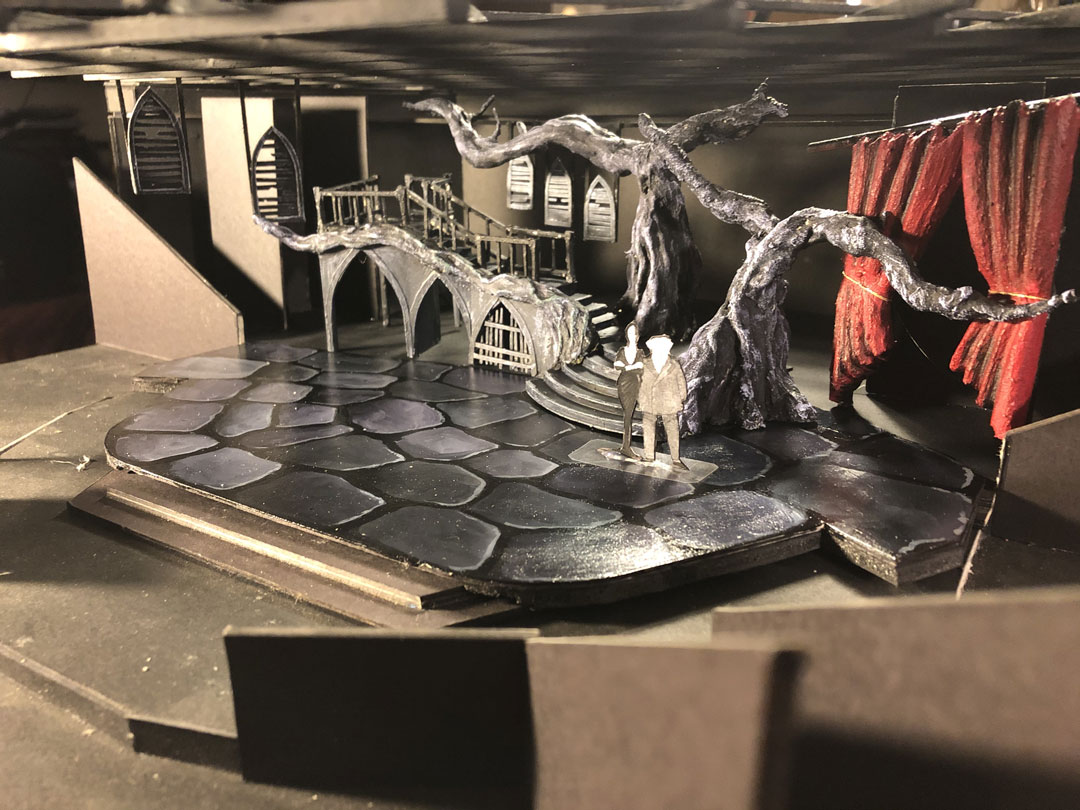 Photo of miniature model of set design for The Addams Family with Gothic staircase, large gnarled tree branches, a hanging red drape, a painted stone floor, and figures of Morticia and Gomez.