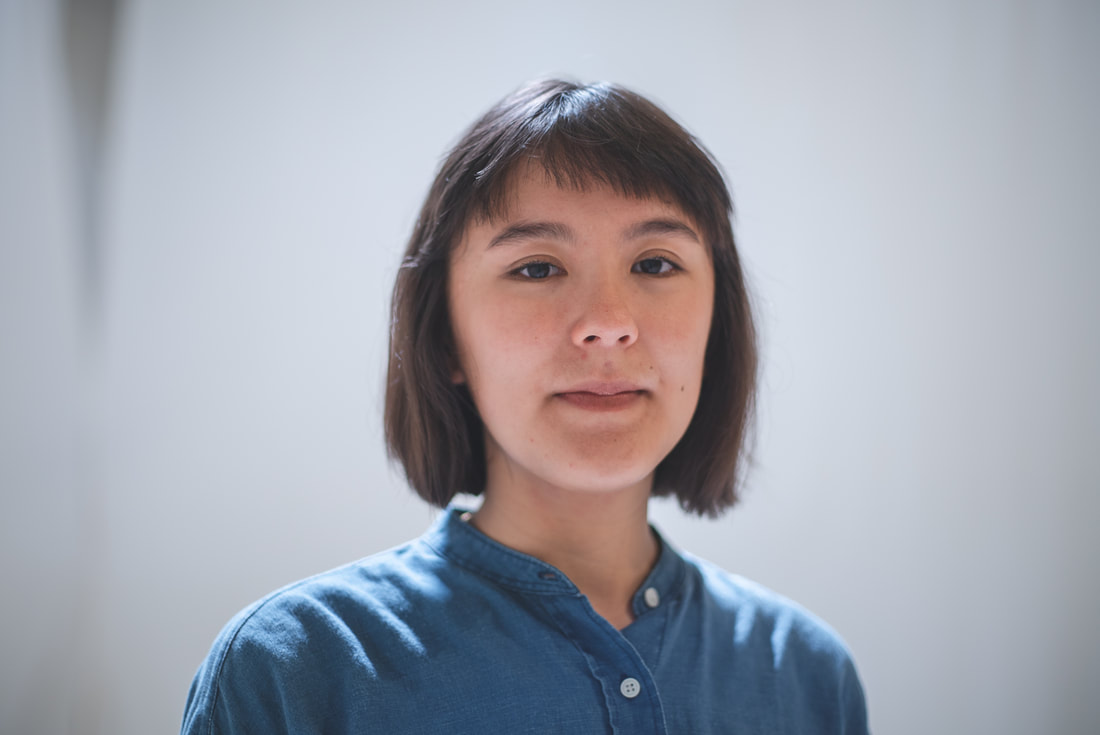 PictureRebecca, a mixed-race woman, has brown chin-length hair with messy bangs and is smiling slightly. She is wearing a blue button-down. She is in front of a blurred white background and sunlight falls onto her shoulders and hair from behind.